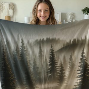 Könnte beinhalten: Eine weiche, graue und weiße Wolldecke mit einem bedruckten Nebelwaldmotiv. Die Decke zeigt eine Silhouette von hohen Bäumen in einem nebelverhangenen Wald.