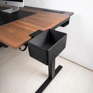 May include: A contemporary wooden desk featuring a black under-desk storage bin. The desk has a dark wood grain finish and a black metal frame. The storage bin is rectangular with a textured surface. A computer monitor is visible.