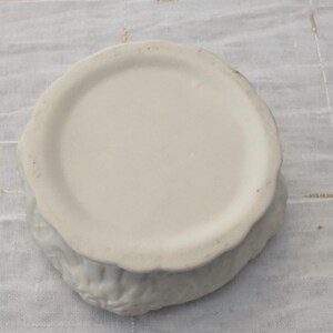 May include: A white ceramic bowl with a scalloped edge. The bowl has a circular base and a textured exterior. The bowl is sitting on a white, textured surface.