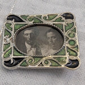 May include: A silver locket with a green and black enamel frame. The locket has a photo of two people inside. The photo is in black and white.