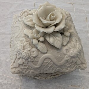 May include: Off-white ceramic jewelry box with a raised floral design. The box is square-shaped with a lid featuring a large rose and smaller floral accents. The sides of the box have a detailed, embossed pattern.