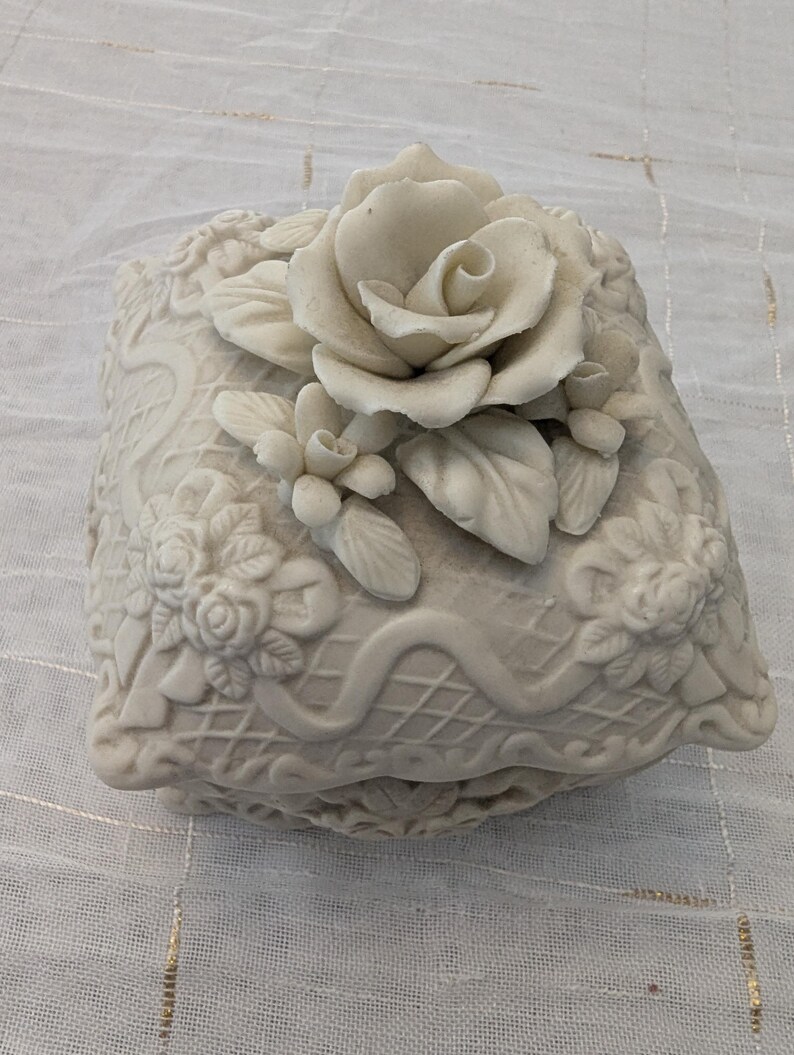 May include: Off-white ceramic jewelry box with a raised floral design. The box is square-shaped with a lid featuring a large rose and smaller floral accents. The sides of the box have a detailed, embossed pattern.