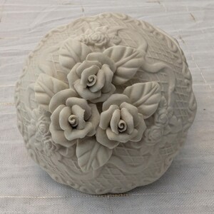 May include: A cream-colored, round decorative container with a lid, adorned with three-dimensional rose and leaf details. The container has a textured surface with additional floral patterns. The roses are in full bloom, adding a touch of elegance.