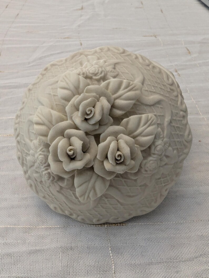 May include: A cream-colored, round decorative container with a lid, adorned with three-dimensional rose and leaf details. The container has a textured surface with additional floral patterns. The roses are in full bloom, adding a touch of elegance.