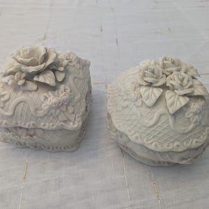 May include: Two off-white decorative trinket boxes. One is square with a rose on top, the other is oval with three roses. Both boxes have floral and lace-like detailing, suggesting a vintage or romantic theme.