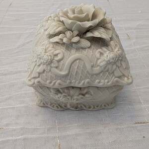 May include: A white ceramic trinket box with a floral design. The lid features a large rose and smaller flowers, with detailed relief patterns. The box has a basket weave design and is set on a white surface.