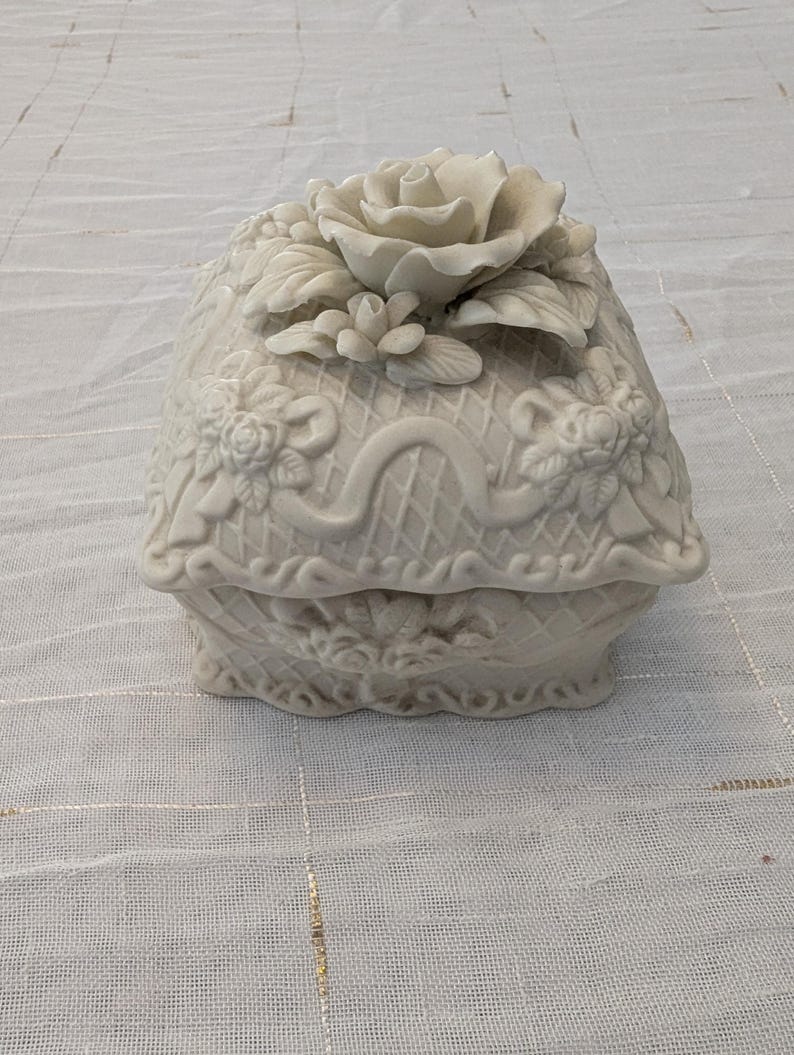 May include: A white ceramic trinket box with a floral design. The lid features a large rose and smaller flowers, with detailed relief patterns. The box has a basket weave design and is set on a white surface.