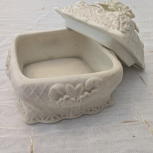 May include: Off-white ceramic jewelry box with a square base and a decorative lid. The lid features floral and lattice designs. The box has a textured surface and is open, revealing the interior.