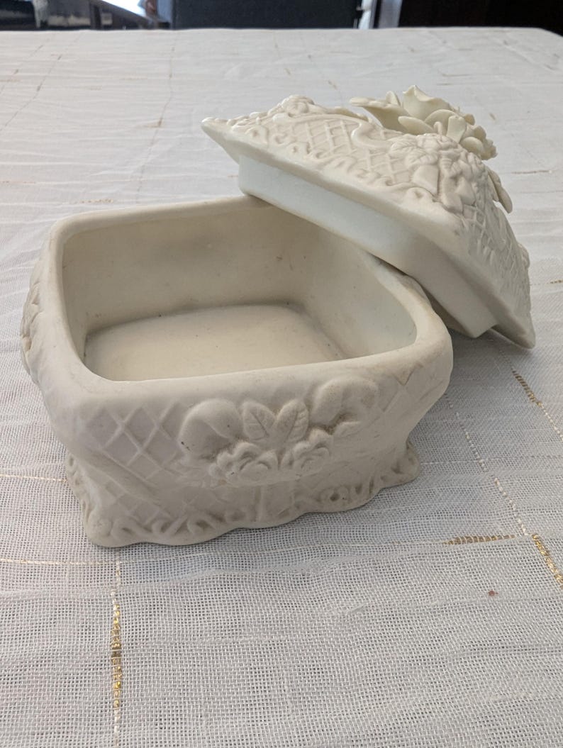 May include: Off-white ceramic jewelry box with a square base and a decorative lid. The lid features floral and lattice designs. The box has a textured surface and is open, revealing the interior.