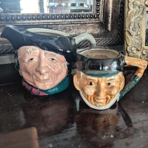 May include: Two ceramic mugs shaped like people's heads. The mugs are painted to look like men wearing hats. One mug is wearing a black hat and the other is wearing a brown hat. Both mugs have facial features and are painted in realistic colours.