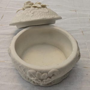 May include: An open, off-white ceramic trinket box with a lid. The lid is decorated with a floral design and a cluster of flowers. The box has a round shape with floral and leaf detailing around the base.