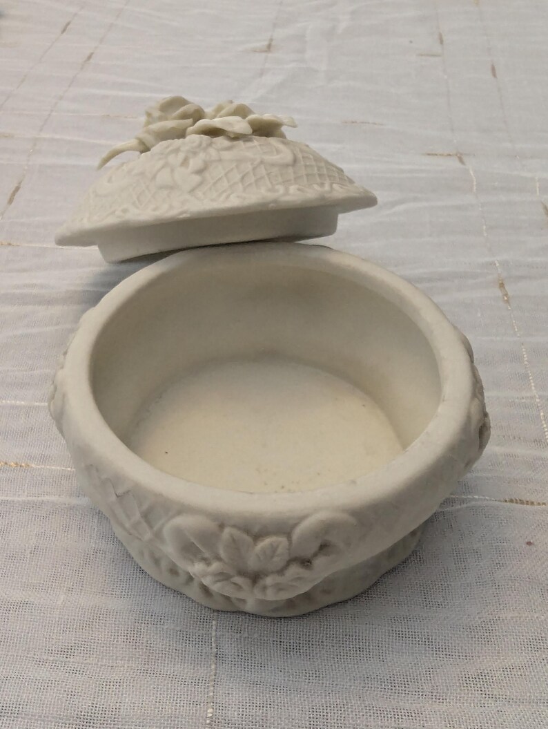 May include: An open, off-white ceramic trinket box with a lid. The lid is decorated with a floral design and a cluster of flowers. The box has a round shape with floral and leaf detailing around the base.