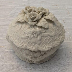 May include: A white, ceramic trinket box with a lid, adorned with sculpted roses and leaves. The box features a basketweave design and ribbon detailing, creating a vintage aesthetic. The box is round and sits on a white surface.