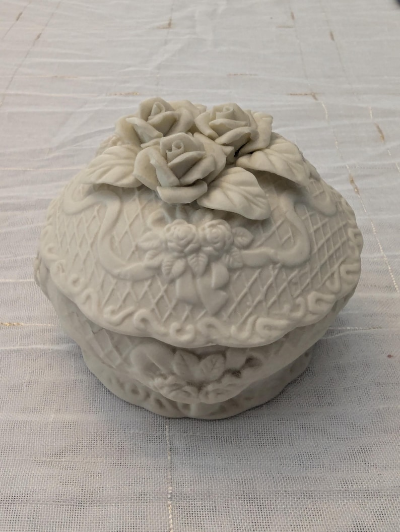 May include: A white, ceramic trinket box with a lid, adorned with sculpted roses and leaves. The box features a basketweave design and ribbon detailing, creating a vintage aesthetic. The box is round and sits on a white surface.