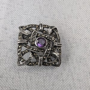 May include: A silver brooch with a square design featuring a purple gemstone surrounded by intricate silver filigree.