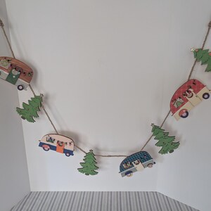 May include: A decorative garland with wooden campervan and Christmas tree cutouts. The campervans are painted in red, blue, and brown, and are connected by twine. The green trees add a festive touch to the holiday decoration.