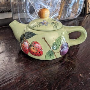 Pode incluir: Bule pequeno de cerâmica verde claro com tampa. Decorado com ilustrações de morangos, ameixas e folhas. A tampa tem um pequeno botão laranja. O bule está sobre uma superfície de madeira escura.