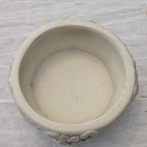 May include: A small, off-white ceramic bowl with a round shape and a slightly textured surface. The bowl has a wide opening and a shallow interior. Decorative floral patterns are visible on the exterior. The bowl is likely used for decorative purposes.