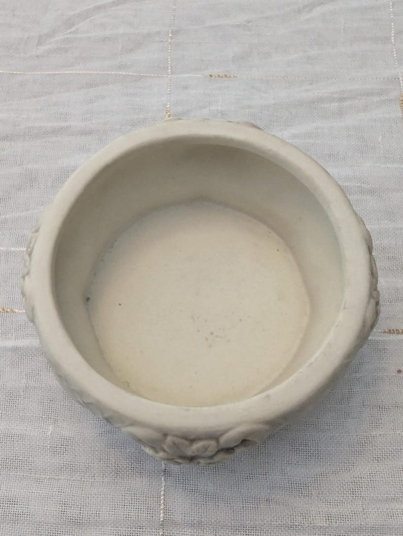 May include: A small, off-white ceramic bowl with a round shape and a slightly textured surface. The bowl has a wide opening and a shallow interior. Decorative floral patterns are visible on the exterior. The bowl is likely used for decorative purposes.