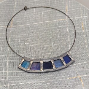 May include: A silver necklace with a stained glass pendant featuring a gradient of blue and purple colors. The pendant is shaped like a crescent moon and is made of six individual pieces of glass.