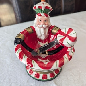 May include: A ceramic Santa Claus dish with a candy cane design. The dish has a small, stainless steel spoon inside. The Santa Claus is wearing a red and white hat with green trim.