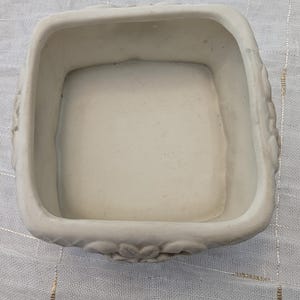 May include: A square, off-white ceramic container with rounded corners. The rim features a floral design. The interior is smooth and the container is empty. The container is likely for decorative or functional use.