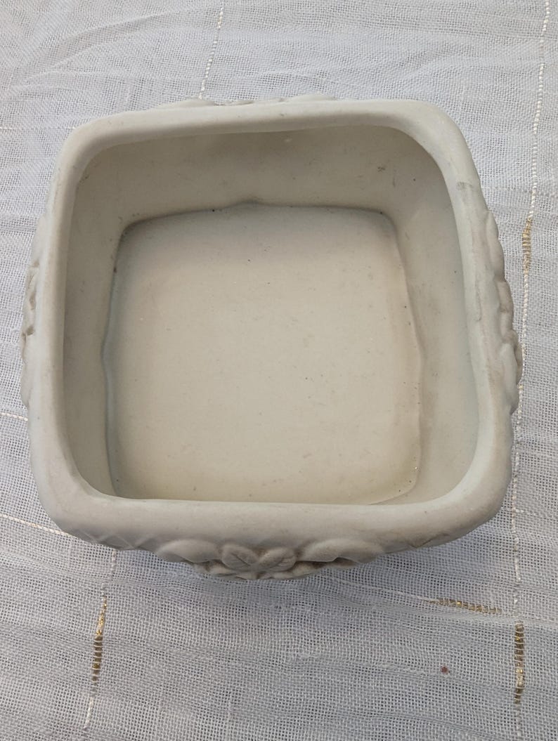 May include: A square, off-white ceramic container with rounded corners. The rim features a floral design. The interior is smooth and the container is empty. The container is likely for decorative or functional use.