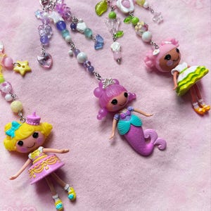 May include: Four colorful keychains featuring small dolls and beaded accents. The dolls include a mermaid, a princess, and two other characters, each with unique outfits and hairstyles. The keychains have silver clasps and are adorned with various beads and charms.
