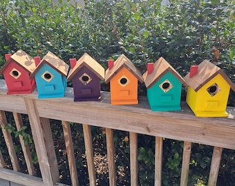 May include: Six colorful birdhouses lined up on a wooden railing. The birdhouses are painted in shades of red, blue, purple, orange, teal, and yellow.