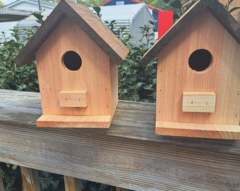 May include: Two wooden birdhouses with brown roofs, each with a round entrance hole and a small rectangular opening below. The birdhouses are sitting on a wooden railing.
