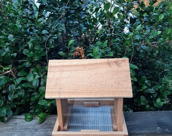Puede incluir: Un comedero para pájaros de madera con un fondo de plástico transparente y un techo de madera con listones. El comedero está sobre una superficie de madera.