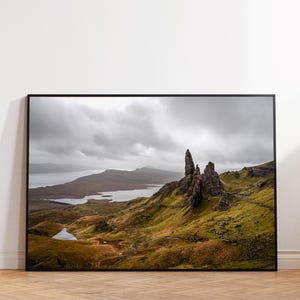 Può includere: Una fotografia paesaggistica incorniciata che ritrae l'Old Man of Storr in Scozia, con formazioni rocciose frastagliate che si ergono su colline verdi, uno specchio d'acqua in lontananza e un cielo nuvoloso dai toni grigi e terrosi.
