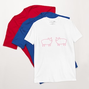 May include: White t-shirt with a pink outline of two sheep facing each other. The t-shirt is layered on top of a blue t-shirt and a red t-shirt.
