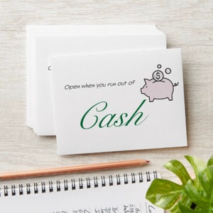 May include: A white card with green text that says "Open when you run out of Cash". A pink piggy bank with a dollar coin on top is illustrated on the card.
