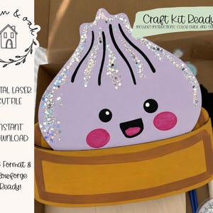 Op de afbeelding: Een lichtpaarse houten uitsparing in de vorm van een dumpling met een lachend gezicht, roze wangen en zilveren glitteraccenten, rustend in een gele houten stoommand-basis. De set bevat de tekst Craft Kit Ready, Digital Laser Cut File en SVG Format.