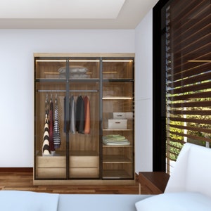 Könnte beinhalten: Ein moderner Holzschrank mit Glastüren, der hängende Kleidung und gefaltete Gegenstände zeigt. Der Schrank hat eine eingebaute Beleuchtung und Schubladen. Der Raum verfügt über ein Bett und ein Fenster mit Holzjalousien.