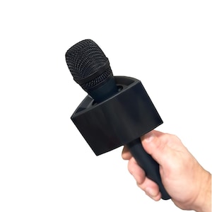 May include: A black microphone with a square base and a black handle is held in a hand. The microphone has a mesh head. The background is white.