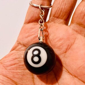 May include: Black and white eight ball keychain. The keychain features a small, round, black ball with a white circle and the number 8 in the center. The keychain is attached to a silver metal ring.