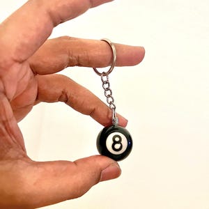 May include: Black and white 8-ball keychain. The keychain features a small, round, black 8-ball with a white number 8. The ball is attached to a silver chain and keyring.