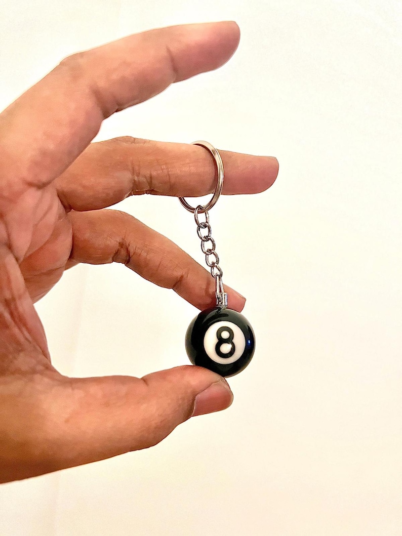 May include: Black and white 8-ball keychain. The keychain features a small, round, black 8-ball with a white number 8. The ball is attached to a silver chain and keyring.