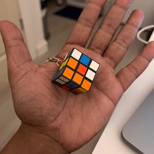 Puede incluir: Un llavero de cubo de Rubik en miniatura. El cubo presenta la cl&aacute;sica disposici&oacute;n de cuadrados de colores: blanco, amarillo, naranja, rojo, azul y verde. El llavero est&aacute; sujeto a una cadena de color plateado.