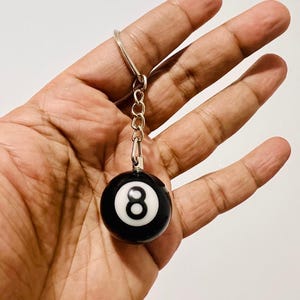 May include: Black and white eight ball keychain. The keychain features a small, round, black ball with a white number eight. The ball is attached to a silver-colored chain and keyring.
