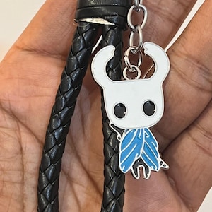 May include: A keychain featuring a white and blue enamel charm of a character with large eyes and horns. The charm is attached to a black braided leather strap with a silver-tone metal keyring.