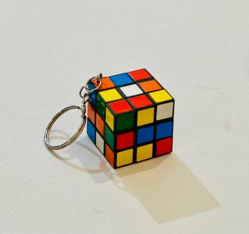 Puede incluir: Un llavero de cubo de Rubik en miniatura. El cubo presenta un dise&ntilde;o cl&aacute;sico con cuadrados coloridos en rojo, azul, amarillo, verde, naranja y blanco. El llavero tiene un anillo de color plateado adjunto al cubo.