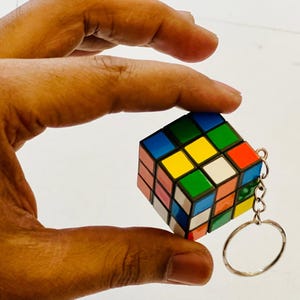 Puede incluir: Un llavero de cubo de Rubik en miniatura con un anillo plateado. El cubo presenta una combinaci&oacute;n de colores cl&aacute;sica: rojo, azul, verde, amarillo, naranja y blanco. El cubo se sostiene en una mano.