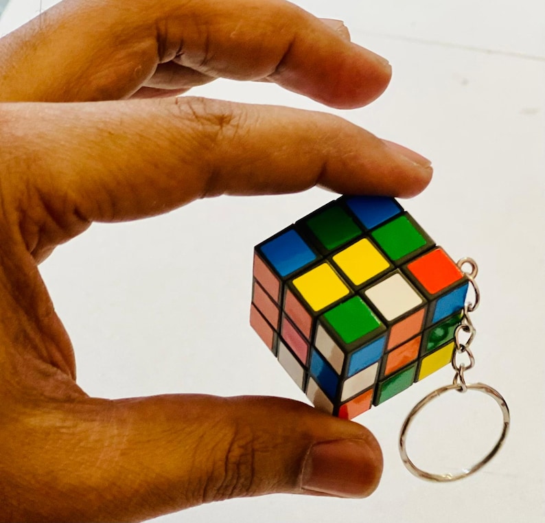 Puede incluir: Un llavero de cubo de Rubik en miniatura con un anillo plateado. El cubo presenta una combinaci&oacute;n de colores cl&aacute;sica: rojo, azul, verde, amarillo, naranja y blanco. El cubo se sostiene en una mano.