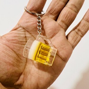 May include: A clear acrylic keychain shaped like a beer mug, filled with yellow liquid. The keychain has a silver chain and ring. The mug has a white foam-like top and a handle. The background is a hand.