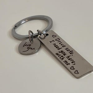 May include: Silver keychain with a rectangular tag and a round charm. The rectangular tag has the words "Drive safe. I need you here, with me" engraved. The round charm says "love you".