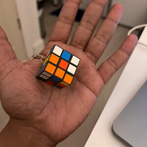 Puede incluir: Un llavero de cubo de Rubik en miniatura. El cubo presenta la disposici&oacute;n cl&aacute;sica de cuadrados de colores: blanco, amarillo, rojo, naranja, azul y verde. El llavero est&aacute; sujeto a una cadena de color plateado.