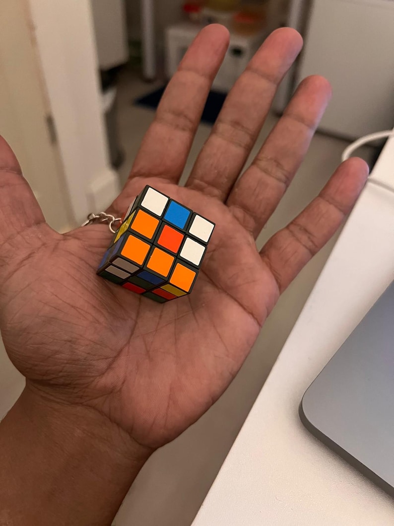 Puede incluir: Un llavero de cubo de Rubik en miniatura. El cubo presenta la disposici&oacute;n cl&aacute;sica de cuadrados de colores: blanco, amarillo, rojo, naranja, azul y verde. El llavero est&aacute; sujeto a una cadena de color plateado.